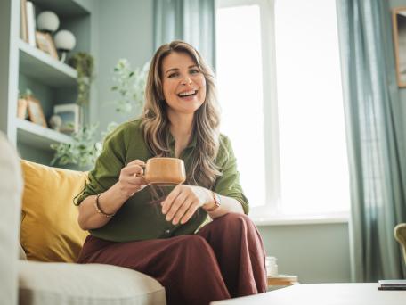 Frau mit Tasse in der Hand sitzt auf einier Couch und lächelt