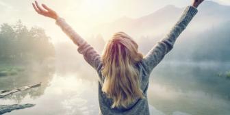 blonde Frau hält diie Arme vor einem See in die Luft